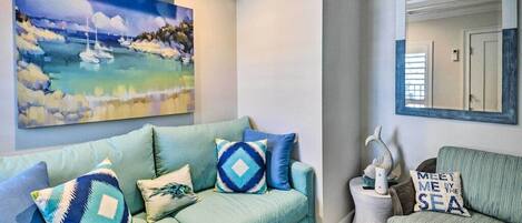 Una sala de estar con un sofá color turquesa, un cuadro de una escena costera y un espejo que refleja una puerta.
