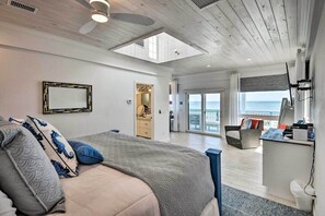 Relax in this serene bedroom with a large bed, ceiling fan, and skylight, offering a view of the ocean from the adjacent balcony.
