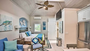 Un dormitorio moderno con cama, sofá azul, ventilador de techo de madera y una zona de cocina con refrigerador.
