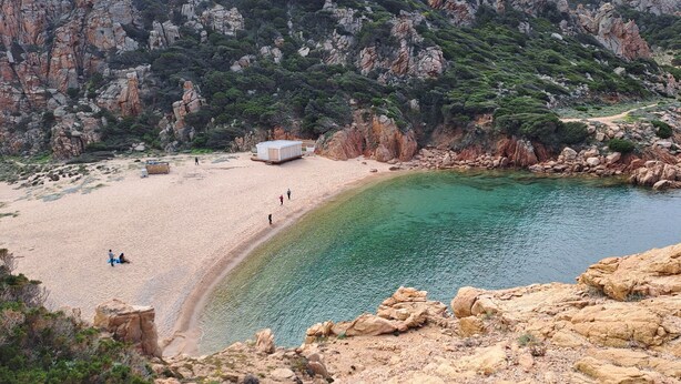A beach with clear water, rocky cliffs, and a small building. - Fantastic view, maximum relax and wellness. (Costa Paradiso)