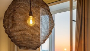 Una mesa de comedor lista para una comida, con una botella de vino, copas y platos. Una lámpara de fibra colgante ilumina el espacio, y se ve una vista del atardecer sobre el agua a través de una ventana abierta.