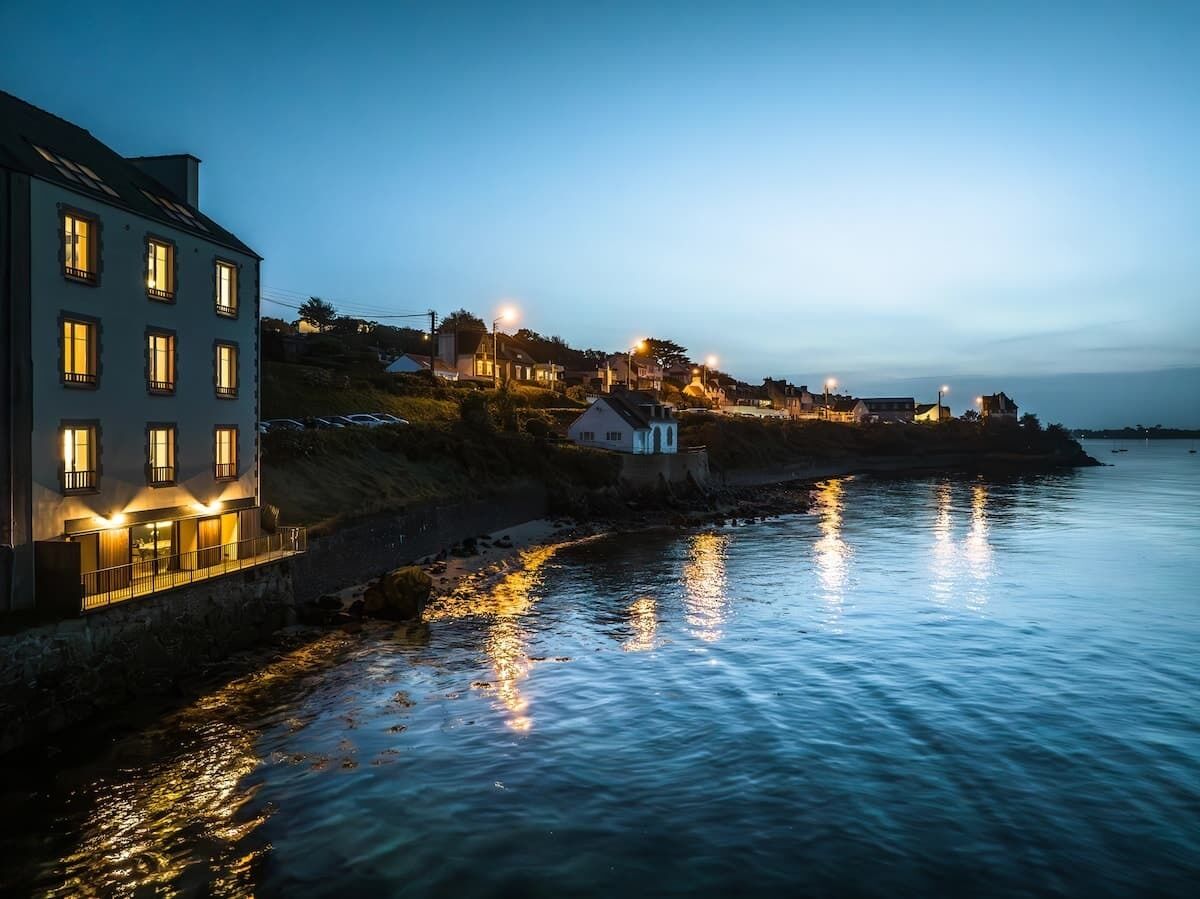 A coastal building with multiple windows and a balcony, situated next to a body of water reflecting lights.