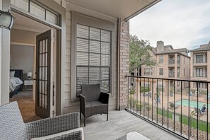 A balcony with wicker furniture, a view of a swimming pool, and a brick building in the background.