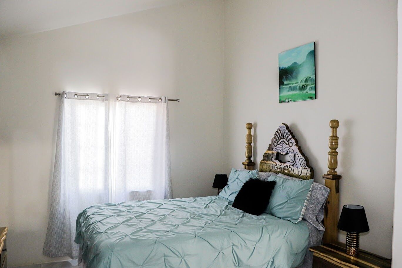 A bedroom with a bed, curtains, a headboard, bedside lamps, and a painting on the wall.