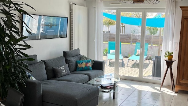 Un salon moderne avec un canapé gris, une table basse en verre et une vue sur une terrasse extérieure dotée d’une piscine et de parasols.