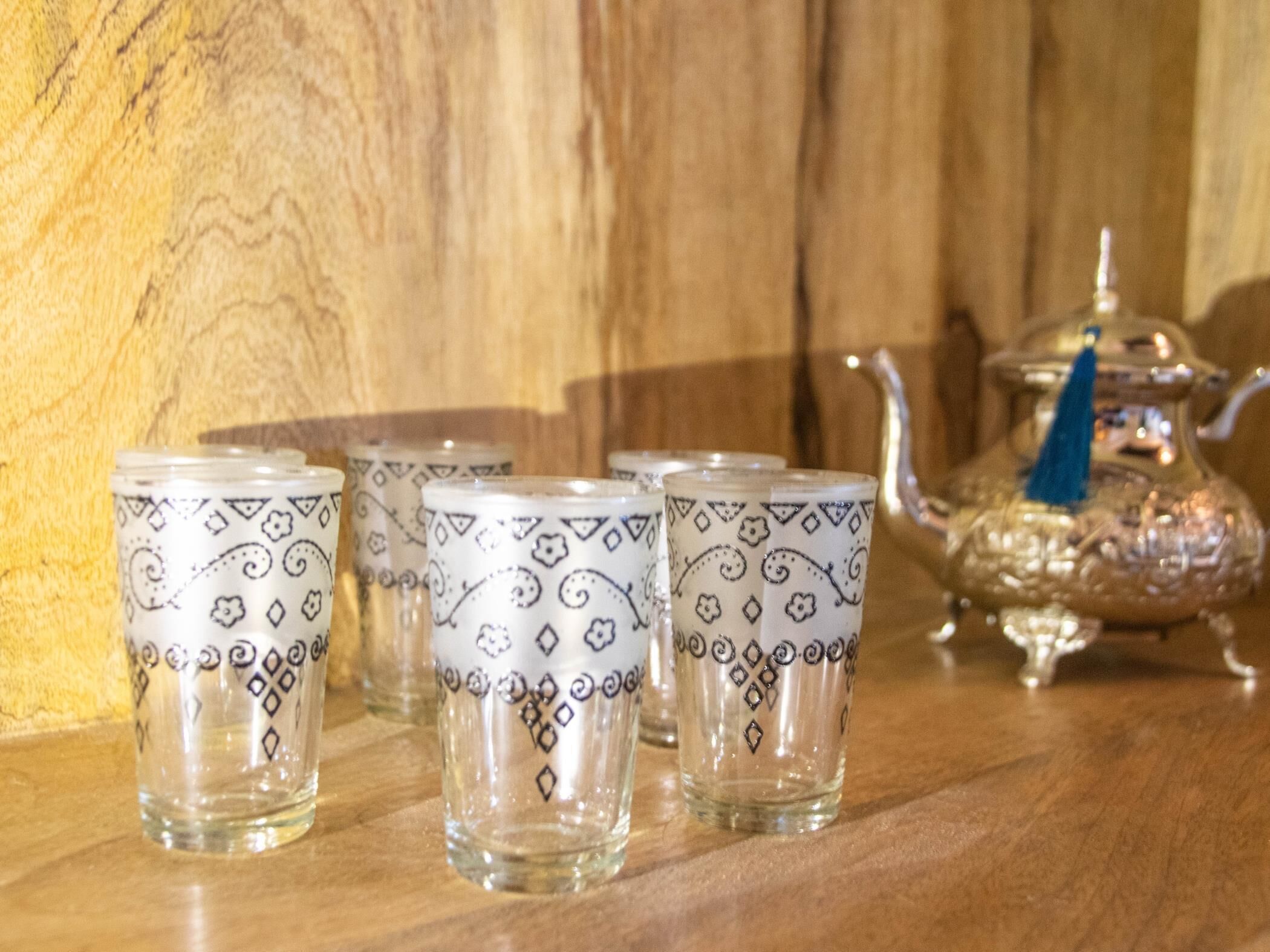Un ensemble de verres en verre transparent ornés de motifs décoratifs, une théière en argent et une pompon bleu sur une surface en bois.