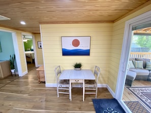 A room with a wooden floor, a white table with two chairs, a painting of a sunset, and a view of a deck through a glass door. - Charming Beach Bungalow: 9 min walk to the Beach (Oak Island)