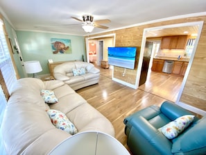 A living room with a beige sectional sofa, a blue armchair, and a flat-screen TV mounted on a wooden wall. - Charming Beach Bungalow: 9 min walk to the Beach (Oak Island)