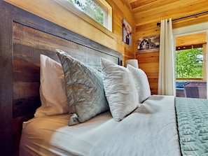 A wooden cabin bedroom with a bed, pillows, and a window with a view of trees.