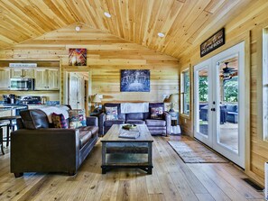A cozy cabin interior with a wooden ceiling, a dining area, a living room with a sofa and coffee table, and a kitchen with a microwave.