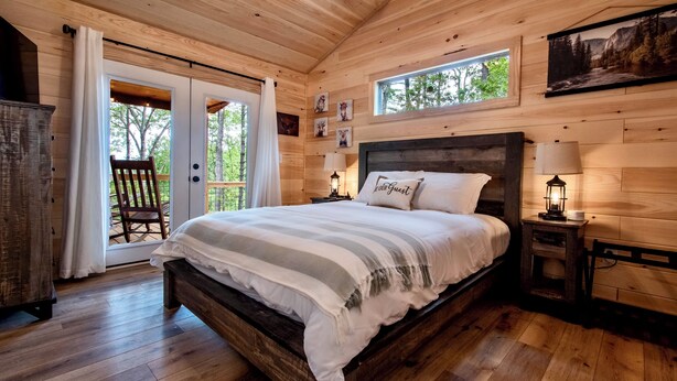 Ein Schlafzimmer in einer Holzhütte mit einem großen Bett, einem Sessel neben der Glasbodentür und einem kleinen Tisch mit einer Lampe.