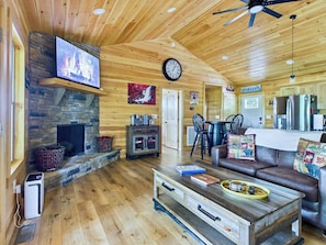 A wooden interior with a stone fireplace, a flat-screen TV, a clock, and a dining area with chairs and a table.