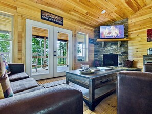 A cozy cabin interior with a stone fireplace, a television mounted above it, and a wooden ceiling.