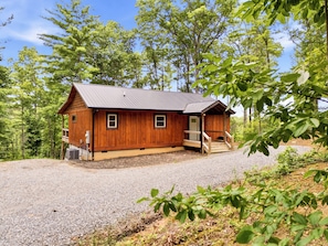 A wooden cabin with a gravel driveway, surrounded by trees.