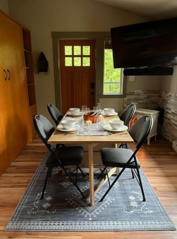 Un comedor con una mesa de madera dispuesta para cuatro personas, rodeada de sillas negras. Una alfombra estampada cubre el suelo, y se ve una puerta de madera con ventanal al fondo.