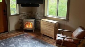 Ein gemütliches Wohnzimmer mit Kamin, einem an der Wand befestigten Fernseher, einem bequemen Ledersessel und einem großen Fenster mit Blick ins Freie.