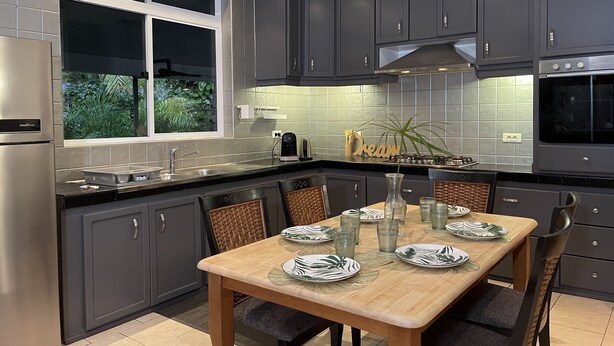 A modern kitchen with dark grey cabinets, a stainless steel refrigerator, a built-in oven, and a dining area with a wooden table and chairs.