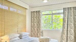 A bedroom with a bed, pillows, a bedside table with a lamp, a window with curtains, and a view of greenery outside.