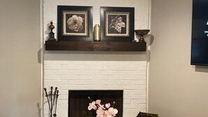 A living room with a white brick fireplace, a wooden coffee table with a vase of flowers, and a television mounted on the wall.