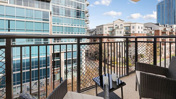 A balcony with wicker furniture and a table, overlooking a cityscape with modern buildings and a clear sky. - Brand New Luxe Apt | 2Mins to Stadium, Free Parking & WiFi, City Views (Charlotte)