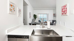 A modern kitchen with a stainless steel sink, white countertops, and a dining area with a table and chairs. - Brand New Luxe Apt | 2Mins to Stadium, Free Parking & WiFi, City Views (Charlotte)