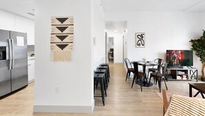 Ett modernt kök med ett kylskåp i rostfritt stål, en matplats med ett träbord och stolar samt ett vardagsrum med en platt-TV och en växt.