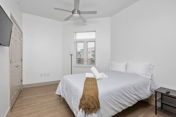 Une chambre à coucher au style minimaliste, comprenant un lit, un ventilateur de plafond, une fenêtre avec des stores et une porte.