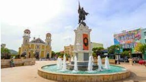 Une place publique avec une fontaine centrale, une statue et une église en arrière-plan.