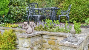 Un jardín con muro de piedra, una estatua de ardilla y un conjunto de mesa y sillas metálicas, todo en medio de abundante vegetación.