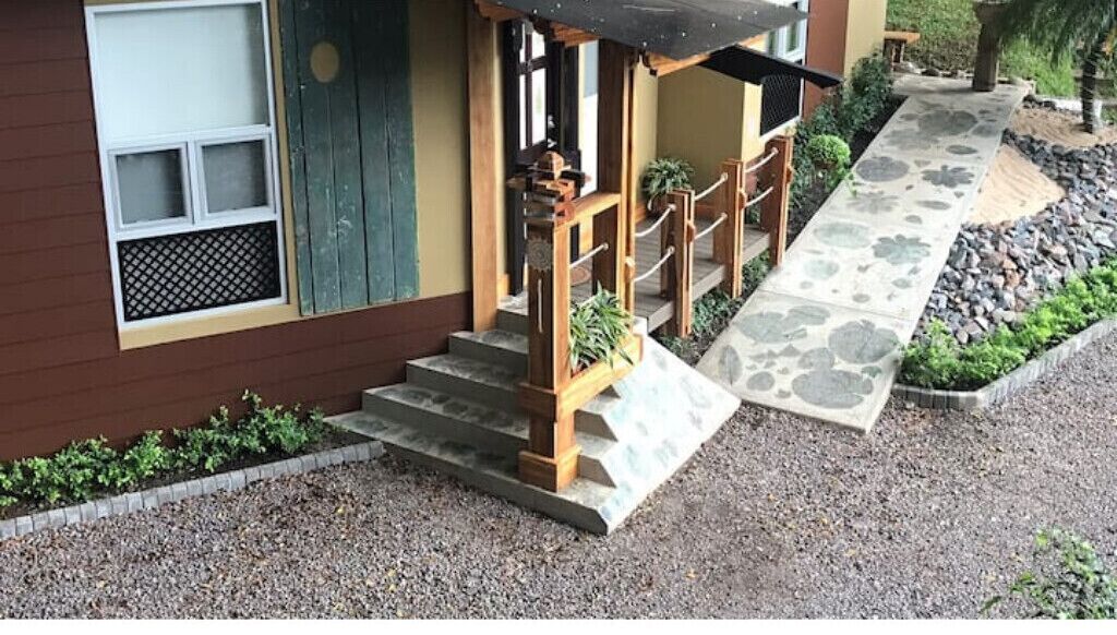 Een gebouw met een houten trap naar een veranda, een klein tuintje met planten en een pad van stenen.