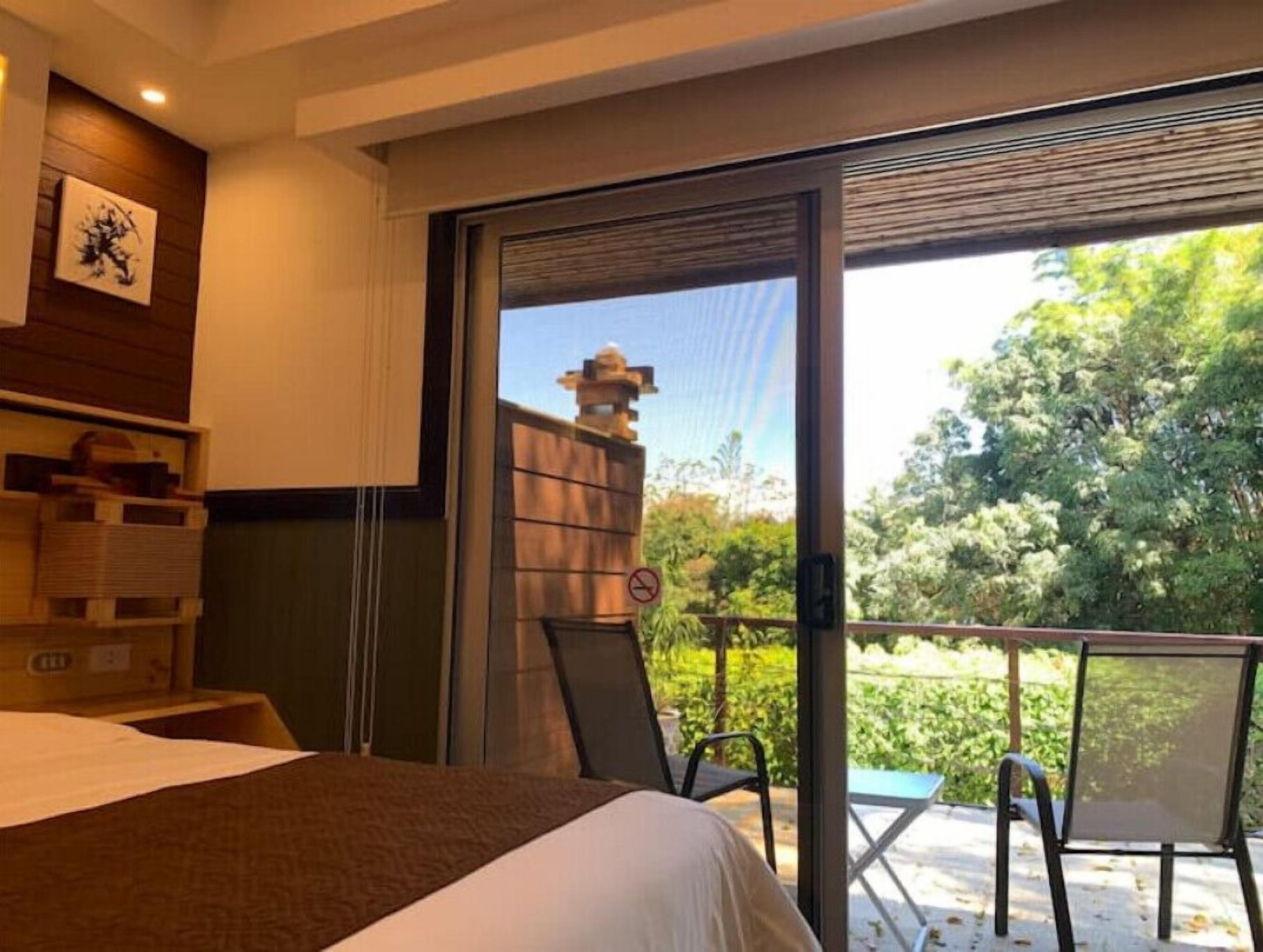 Ein Hotelzimmer mit Bett, Schreibtisch, Stuhl und einem Balkon mit Blick auf Bäume und ein Gebäude.