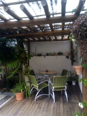 En overdækket terrasse med et rundt bord og fire stole, omgivet af plantekrukker og et træpergola.