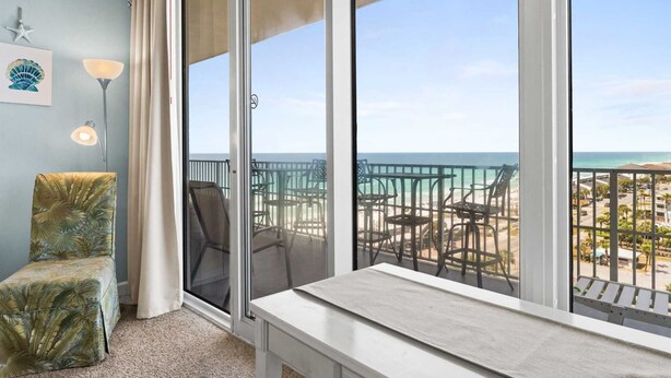 A balcony with a view of the beach and ocean, a chair, a table, and a lamp.