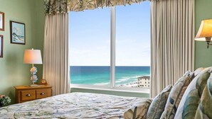 A bedroom with a bed, a nightstand, a lamp, a window with curtains, and a view of the ocean.