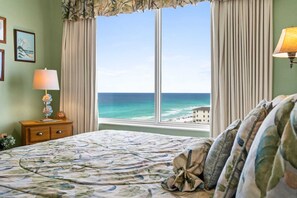 A bedroom with a bed, a nightstand, a lamp, a window with curtains, and a view of the ocean.