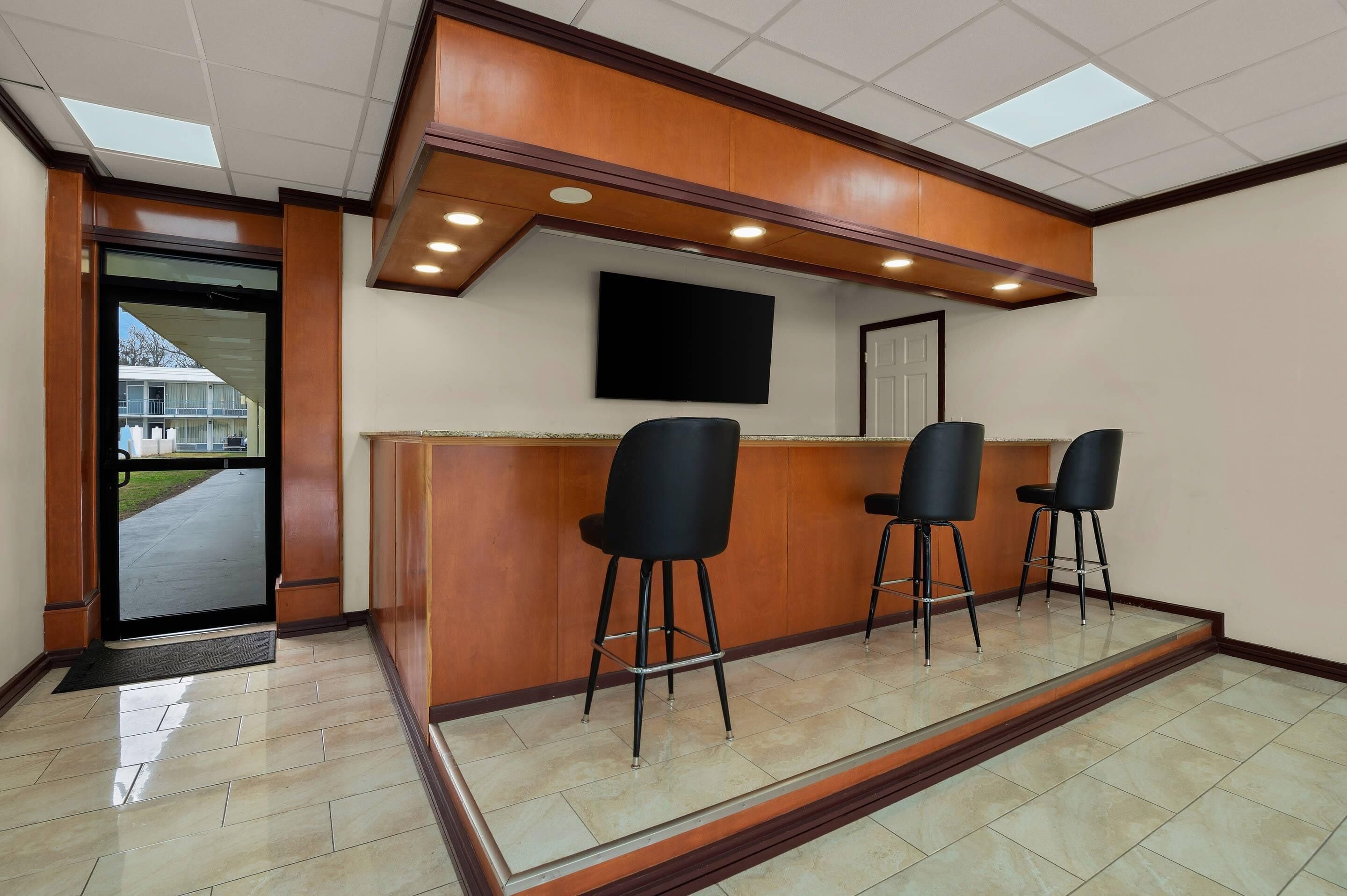 Zona de recepción con mostrador de madera, tres taburetes negros, un televisor de pantalla plana montado en la pared y una puerta que da al exterior.