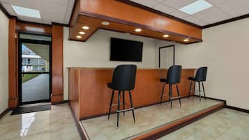 Zona de recepción con mostrador de madera, tres taburetes negros, un televisor de pantalla plana montado en la pared y una puerta que da al exterior.