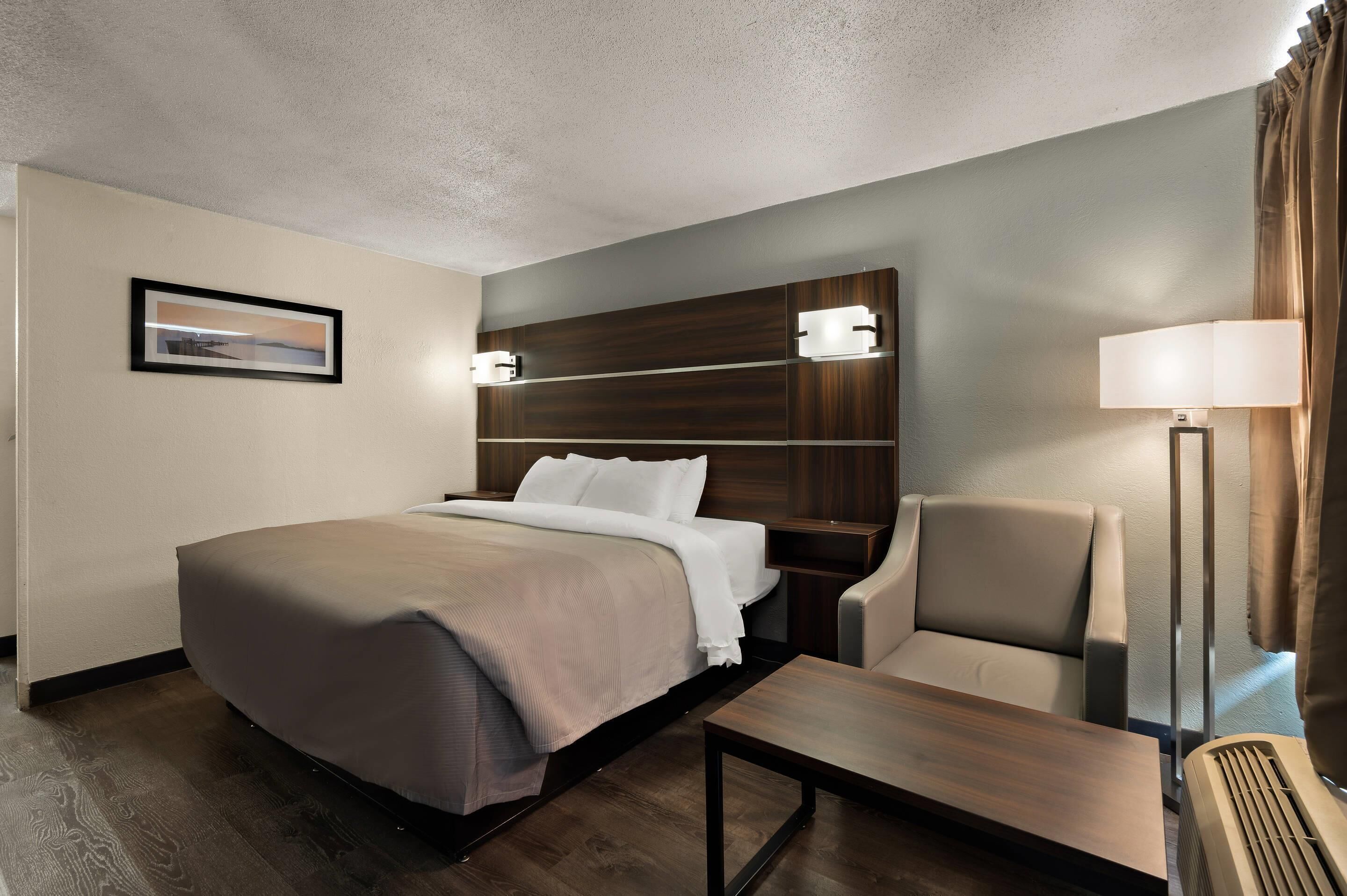 A hotel room with a bed, a chair, a desk, a lamp, and a picture on the wall.
