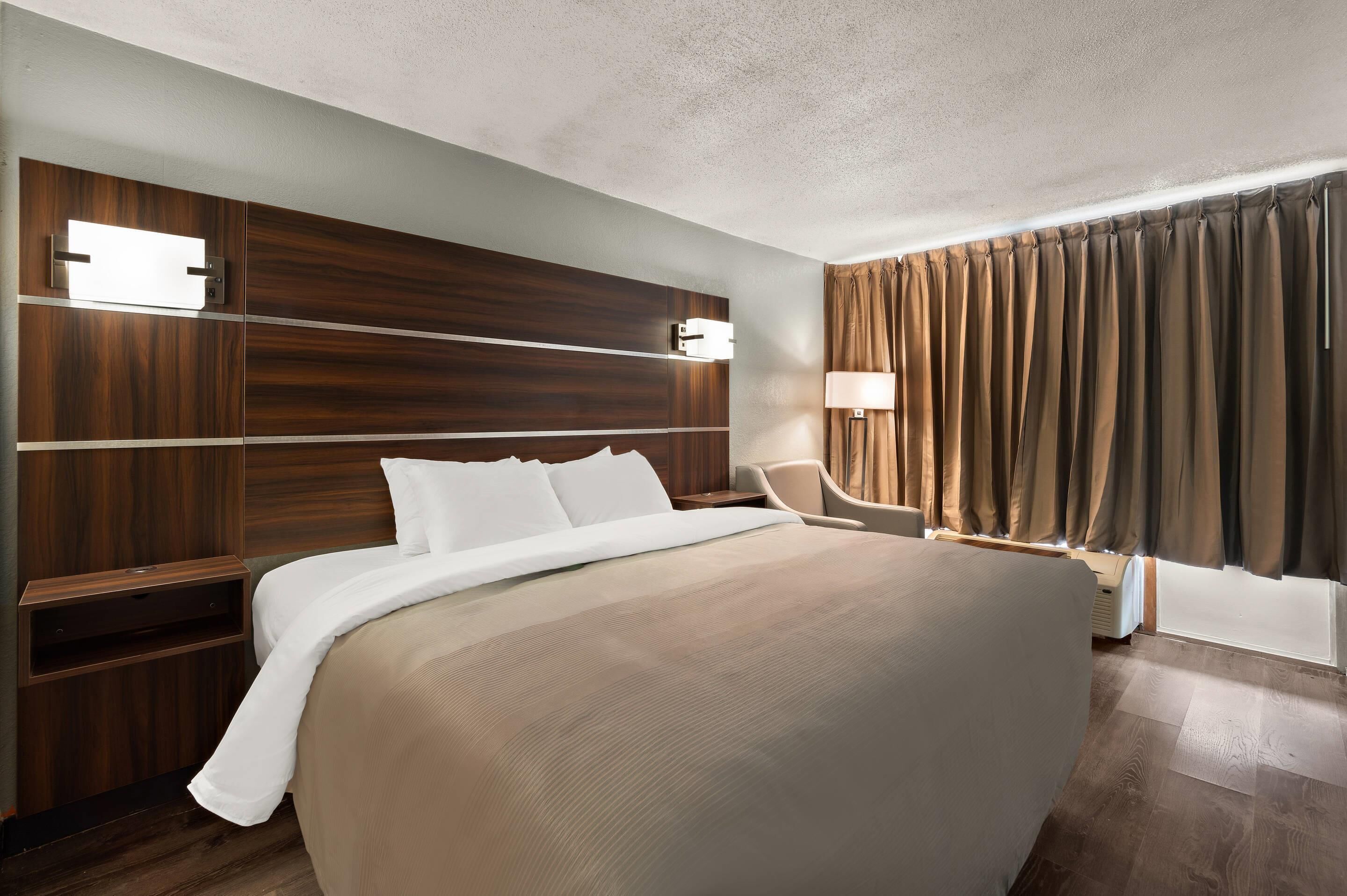 A hotel room with a large bed, wooden headboard, bedside tables, a desk, a chair, a lamp, and a window with curtains.