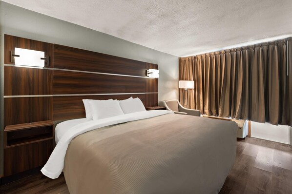 Quarto de hotel com uma cama grande, cabeceira de madeira, mesas de cabeceira, uma escrivaninha, uma cadeira, um abajur e uma janela com cortinas.