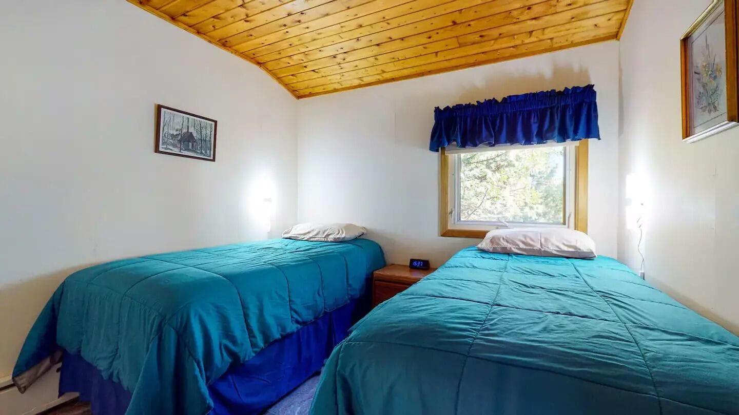 A room with two beds, a window with a blue valance, a wooden ceiling, and a framed picture on the wall.