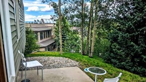 Une terrasse avec deux chaises blanches et une petite table ronde, entourée d’une zone en béton et d’un jardin verdoyant avec des arbres.