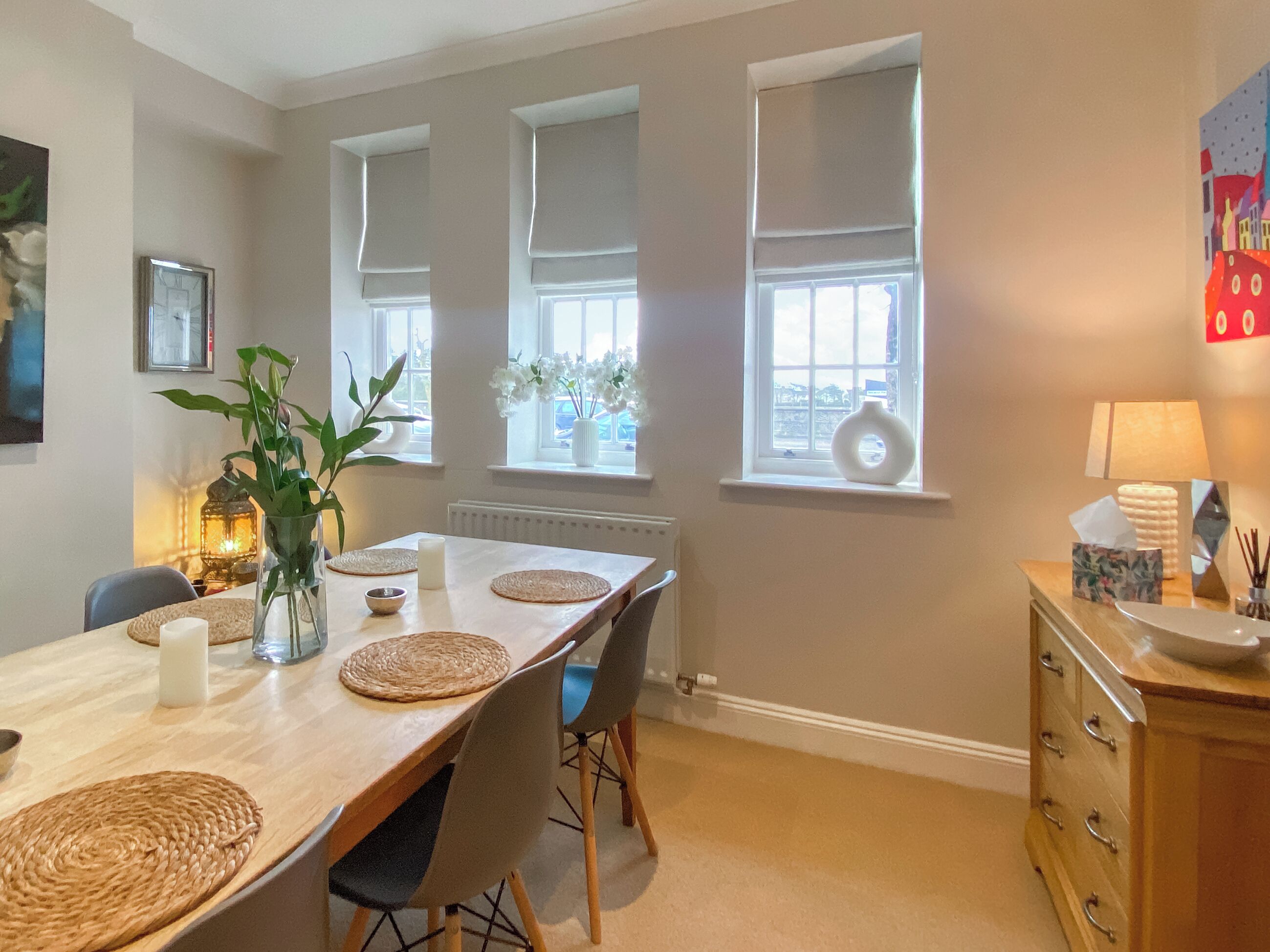 A dining area with a wooden table, chairs, and a vase with flowers. There are windows with blinds, a radiator, and a lamp on a side table.
