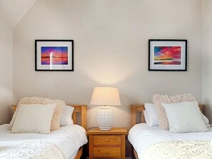Une chambre avec deux lits, chacun doté de draps blancs et d’oreillers moelleux, une table de chevet avec une lampe, et deux tableaux encadrés accrochés au mur.
