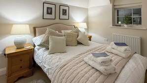 Una cama bien hecha con colcha, almohadas y toallas dobladas. Hay dos lámparas de noche, un mesita auxiliar de madera y una ventana con una planta.