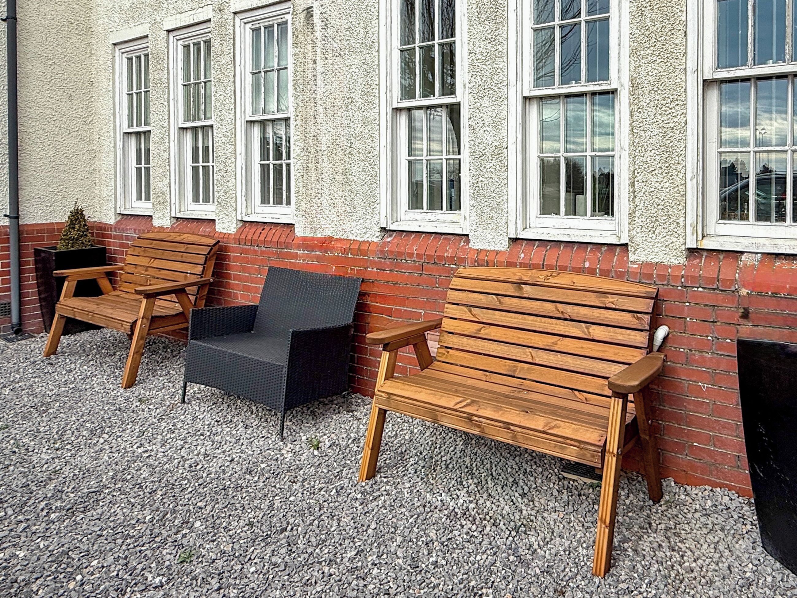 Deux bancs en bois et une chaise noire sont placés à l’extérieur d’un bâtiment doté de fenêtres à cadre blanc et d’un mur en briques rouges.