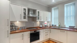 A modern kitchen with white cabinets, a built-in oven, a coffee machine, and a window with blinds. - The Court House - UK43908 (Silloth)