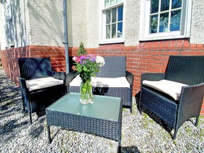 Un espace extérieur aménagé avec des chaises en osier et une table en verre, un vase rempli de fleurs et un mur de briques percé de fenêtres.