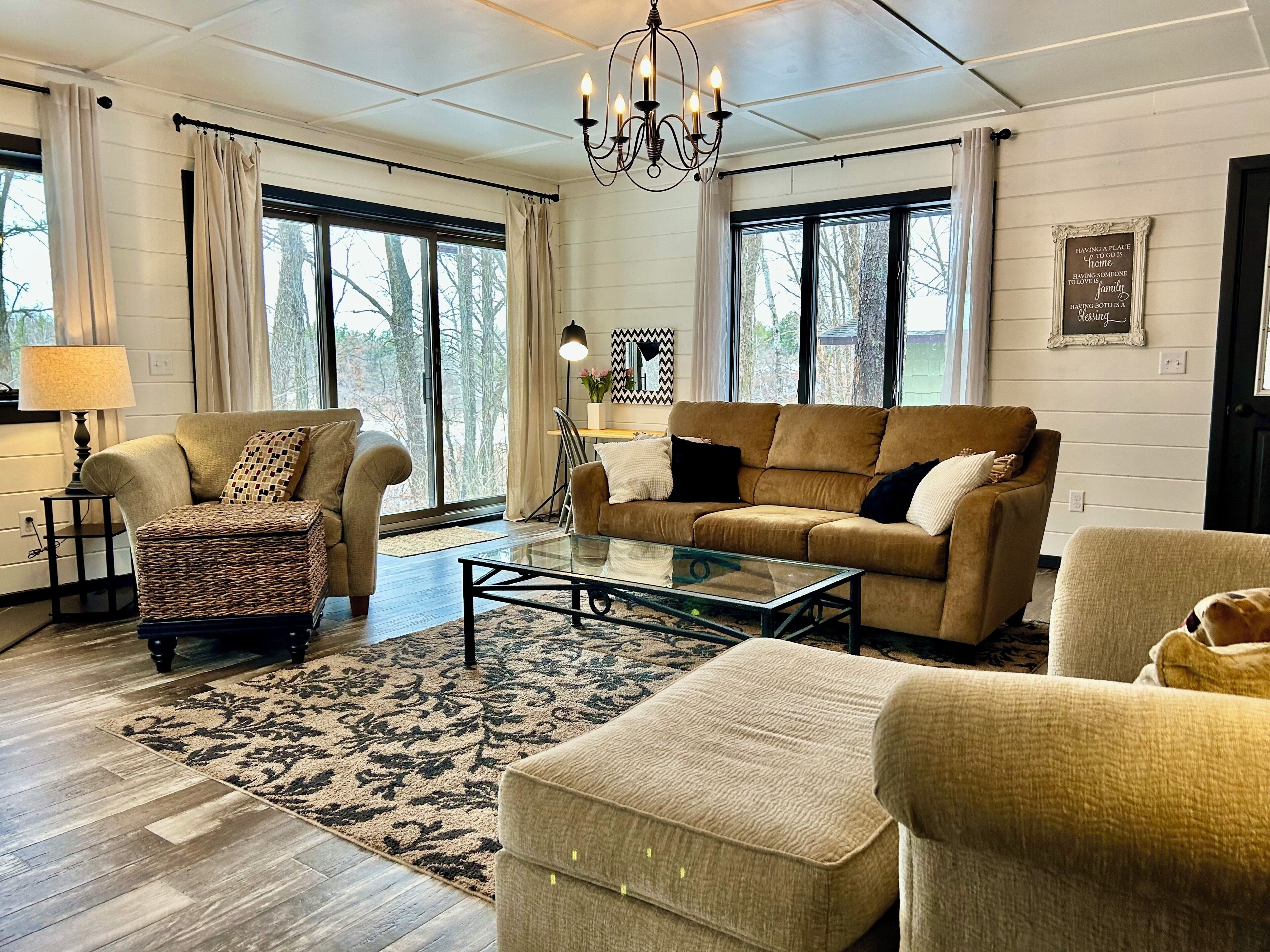 A living room with a sofa, armchair, ottoman, glass coffee table, and a chandelier.