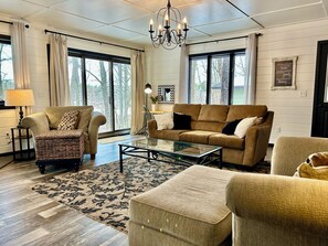 A living room with a sofa, armchair, ottoman, glass coffee table, and a chandelier.